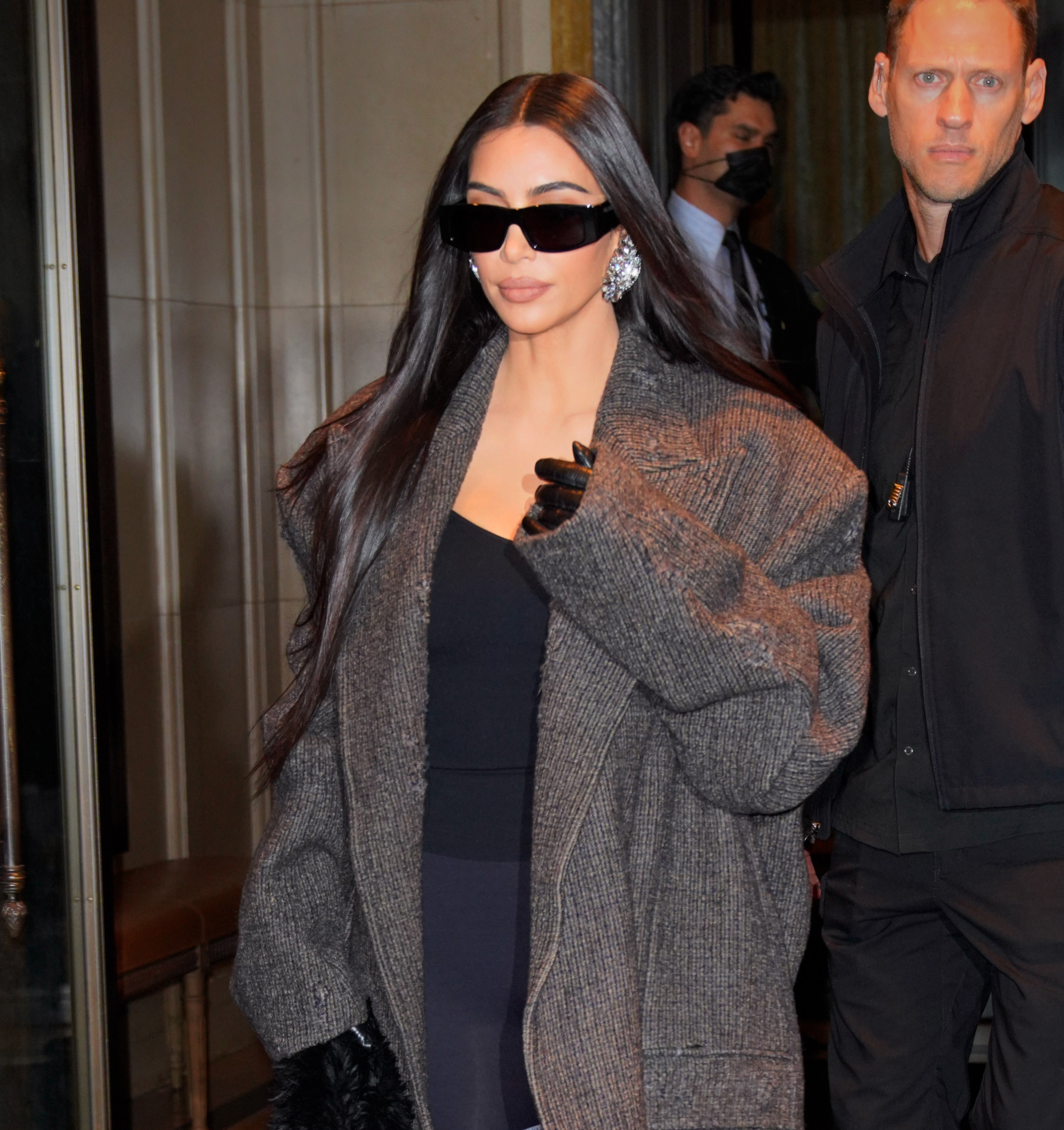 Kim walking out of a building wearing a large coat and sunglasses