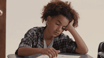 A young girl at a school desk with her head resting on one of her hands and the other taking notes