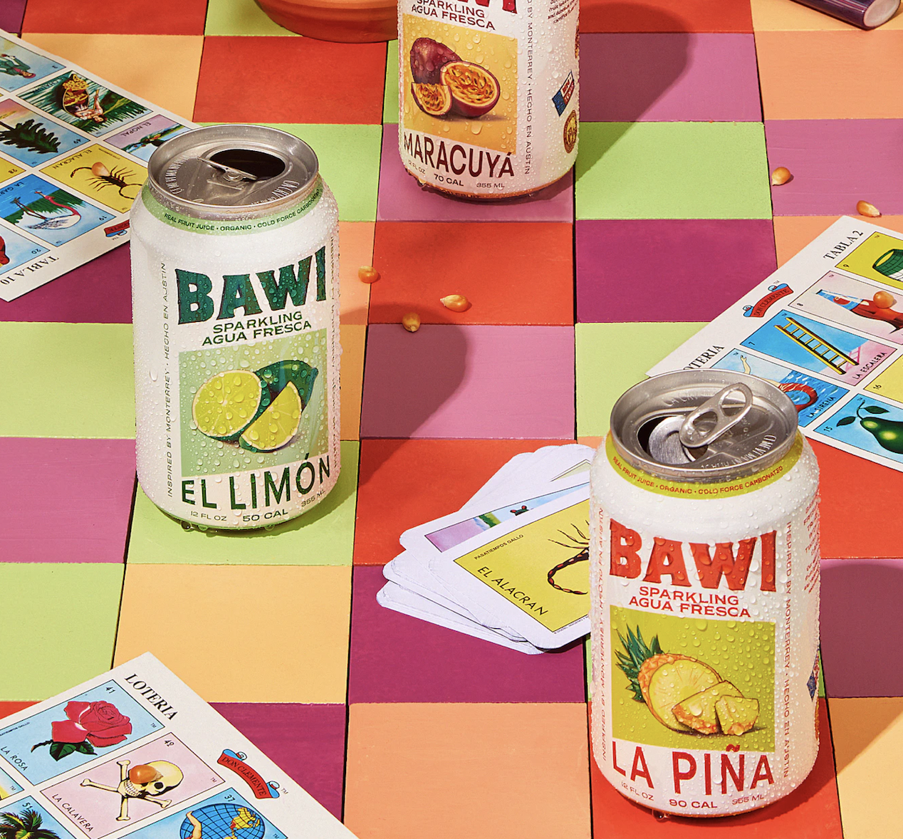 Three cans of agua frescas sitting near Loteria cards on colorful tiles.