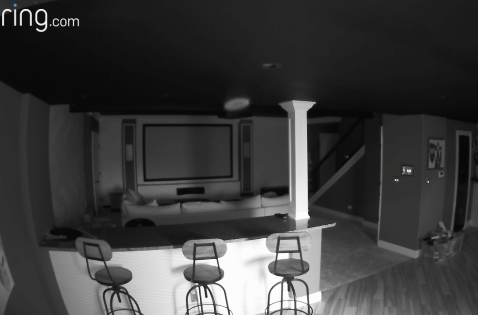 Screenshot of a darkened room with a large TV and three chairs at a bar and stairs in the background