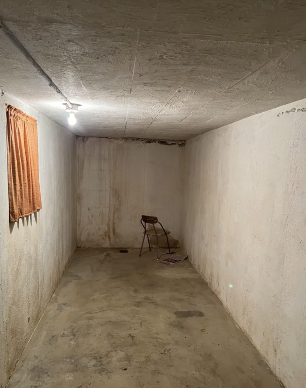 Empty, narrow, windowless room with dirty walls, a single light bulb, and a metal folding chair