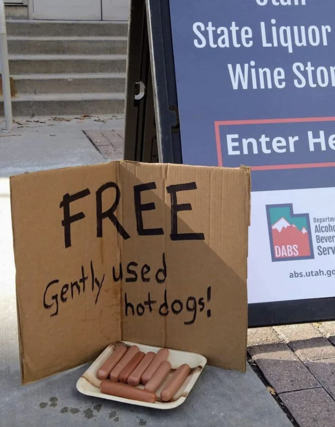 A plate full of hot dogs sits on the sidewalk, and a sign next to it advertises &quot;free gently used hot dogs&quot;