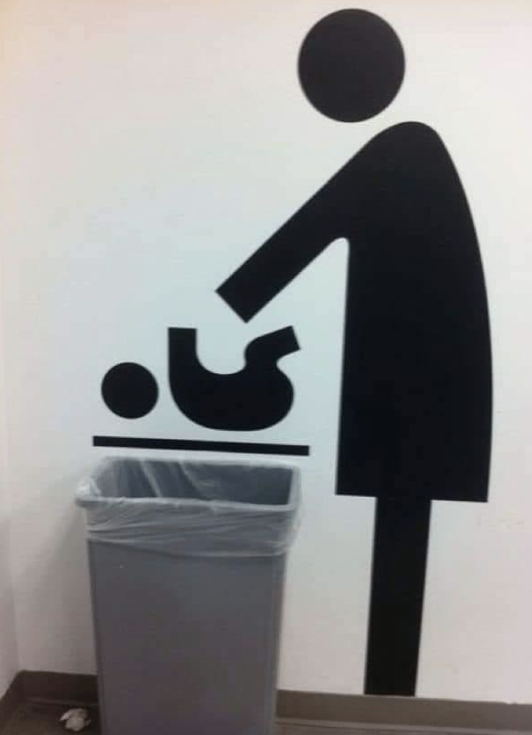 A sign on the wall features a graphic representing a baby changing station, but someone moved a trash can so it looks like the graphic is throwing the baby away&quot;
