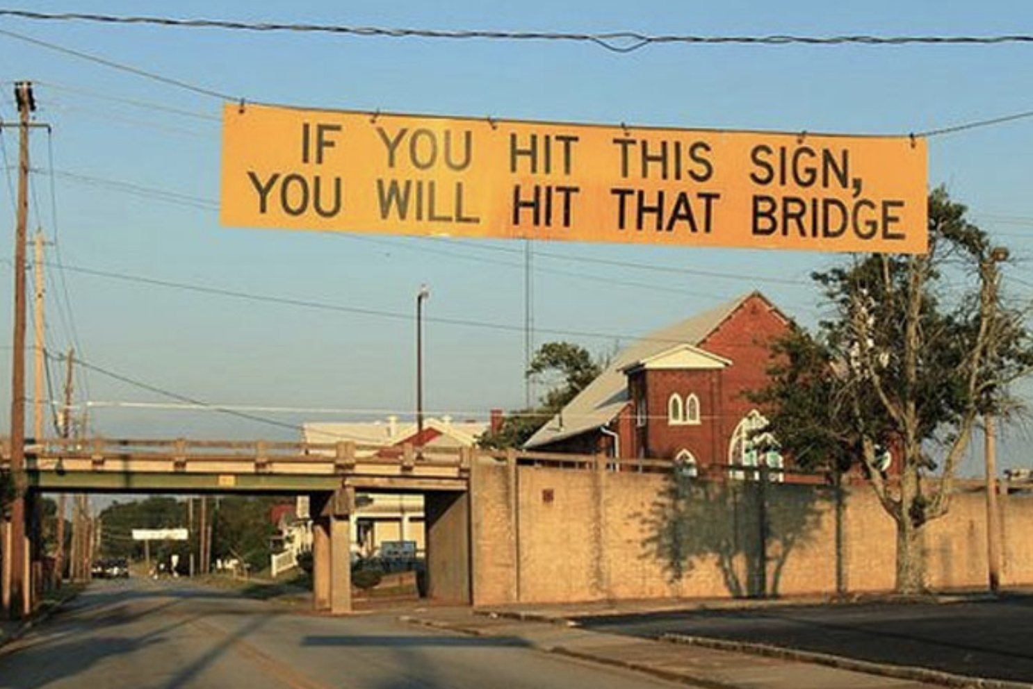 A sign hanging over a street in front of an overpass says &quot;if you hit this sign, you will hit that bridge&quot;