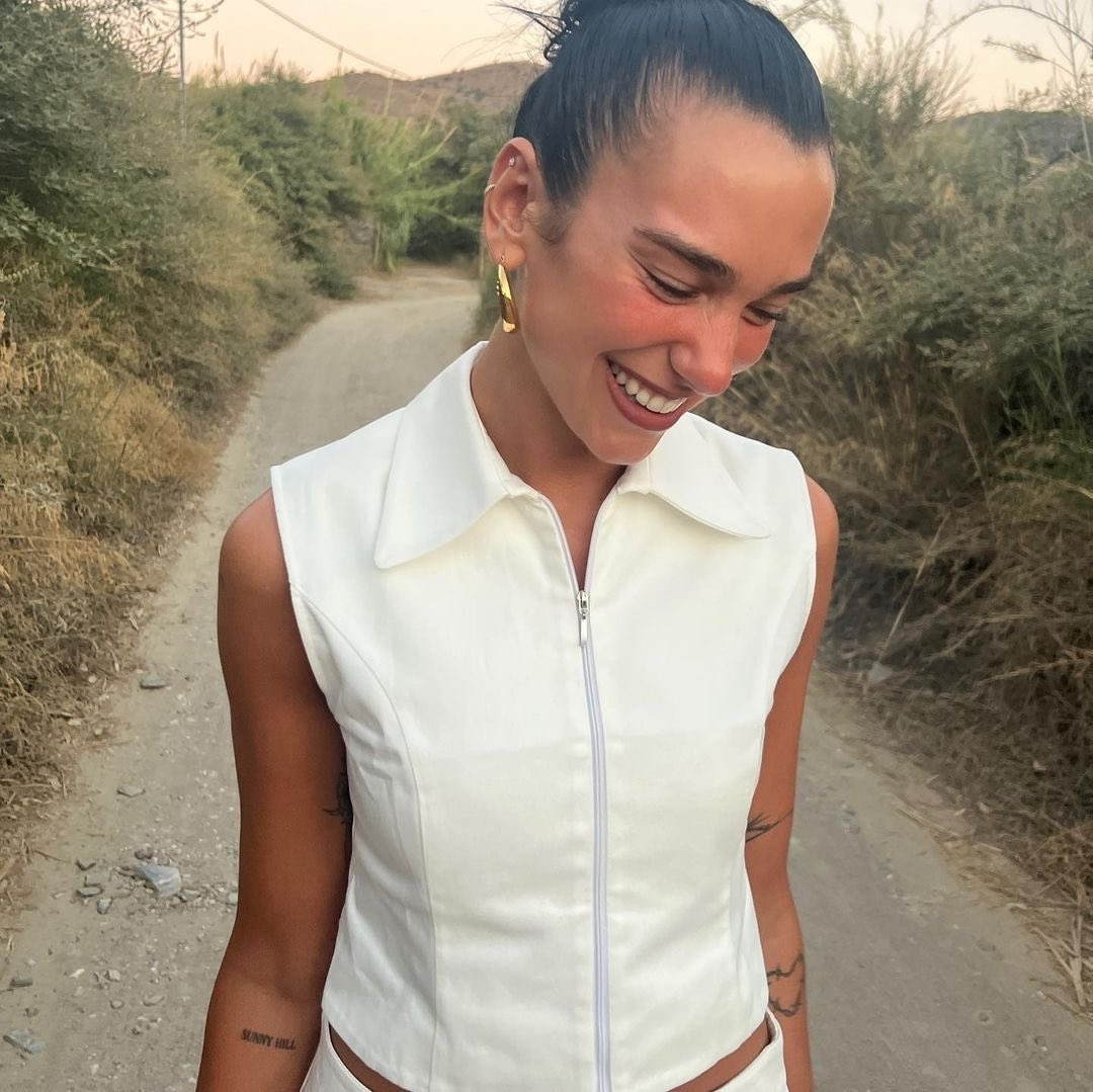 Dua LIpa smiles looking down with her hair back and a tan.