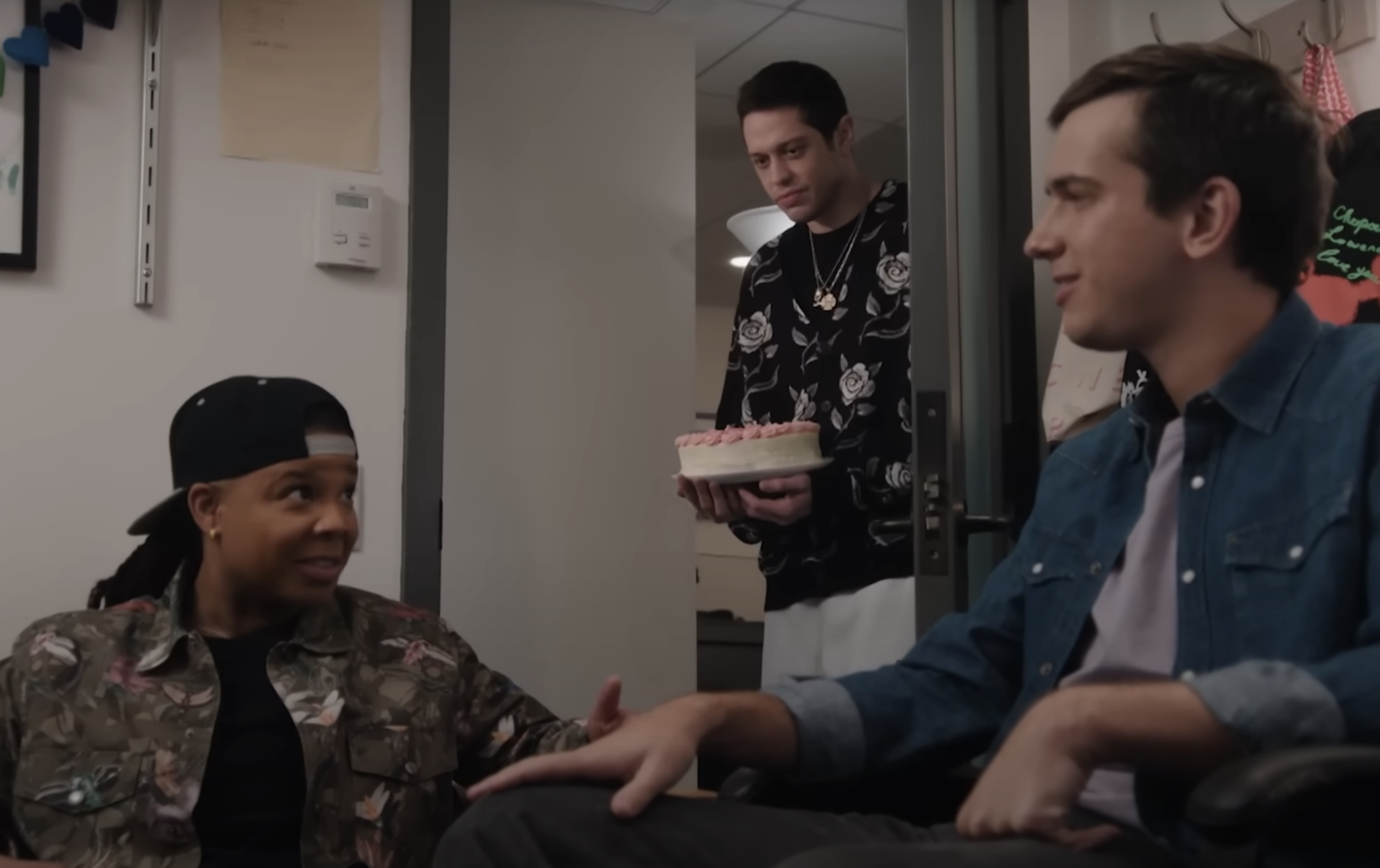 Pete holding a cake in a doorway and looking on at two people talking