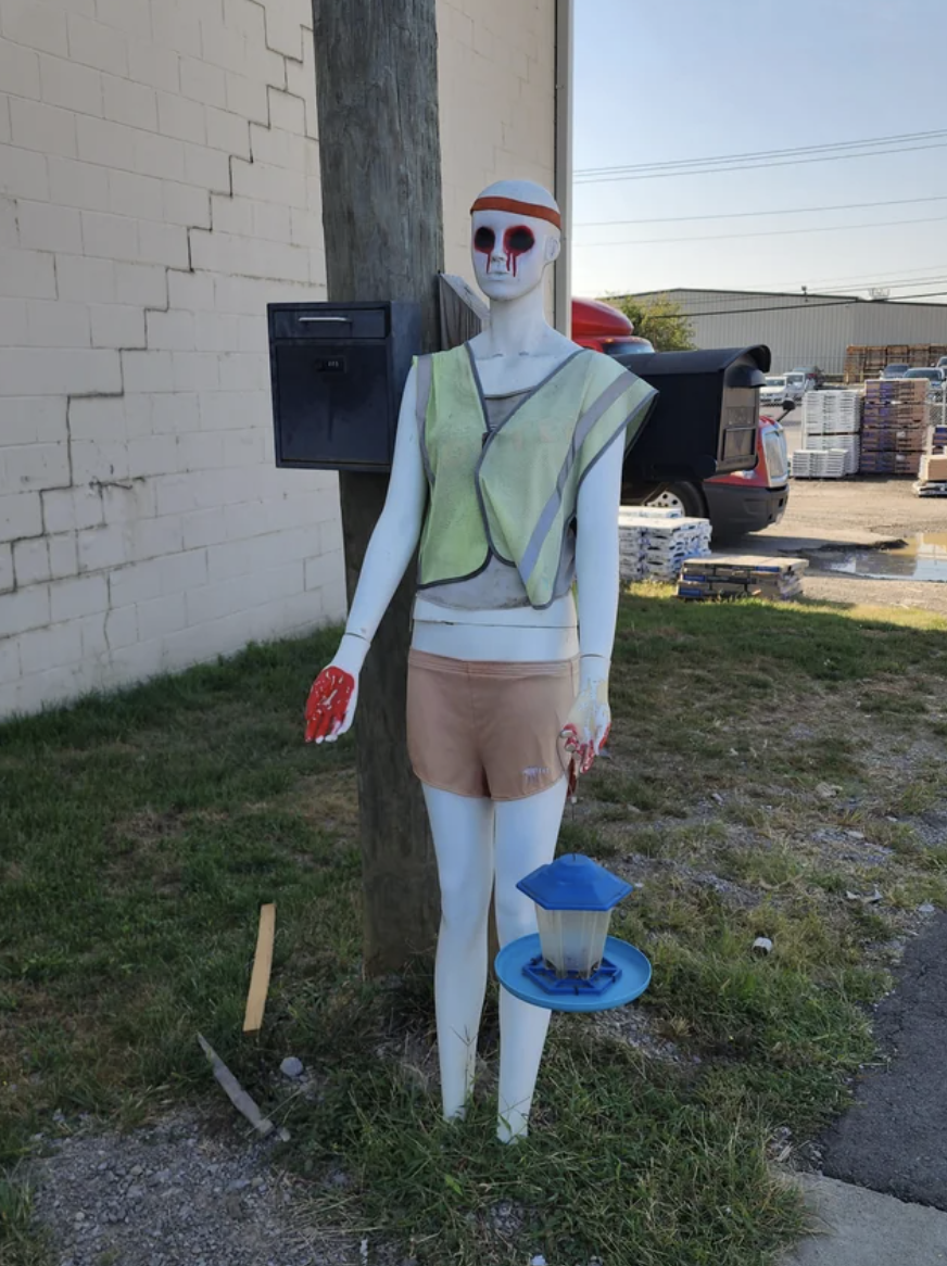 A mannequin with gouged-out eyes and what looks like dripping blood around the sockets standing by a wooden beam on the grass