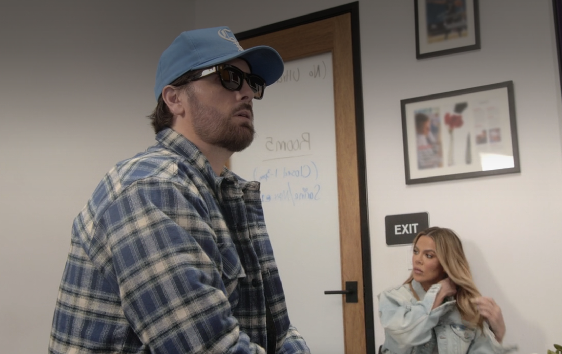 Close-up of Scott in a plaid jacket, cap, and sunglasses with Khloé sitting behind him
