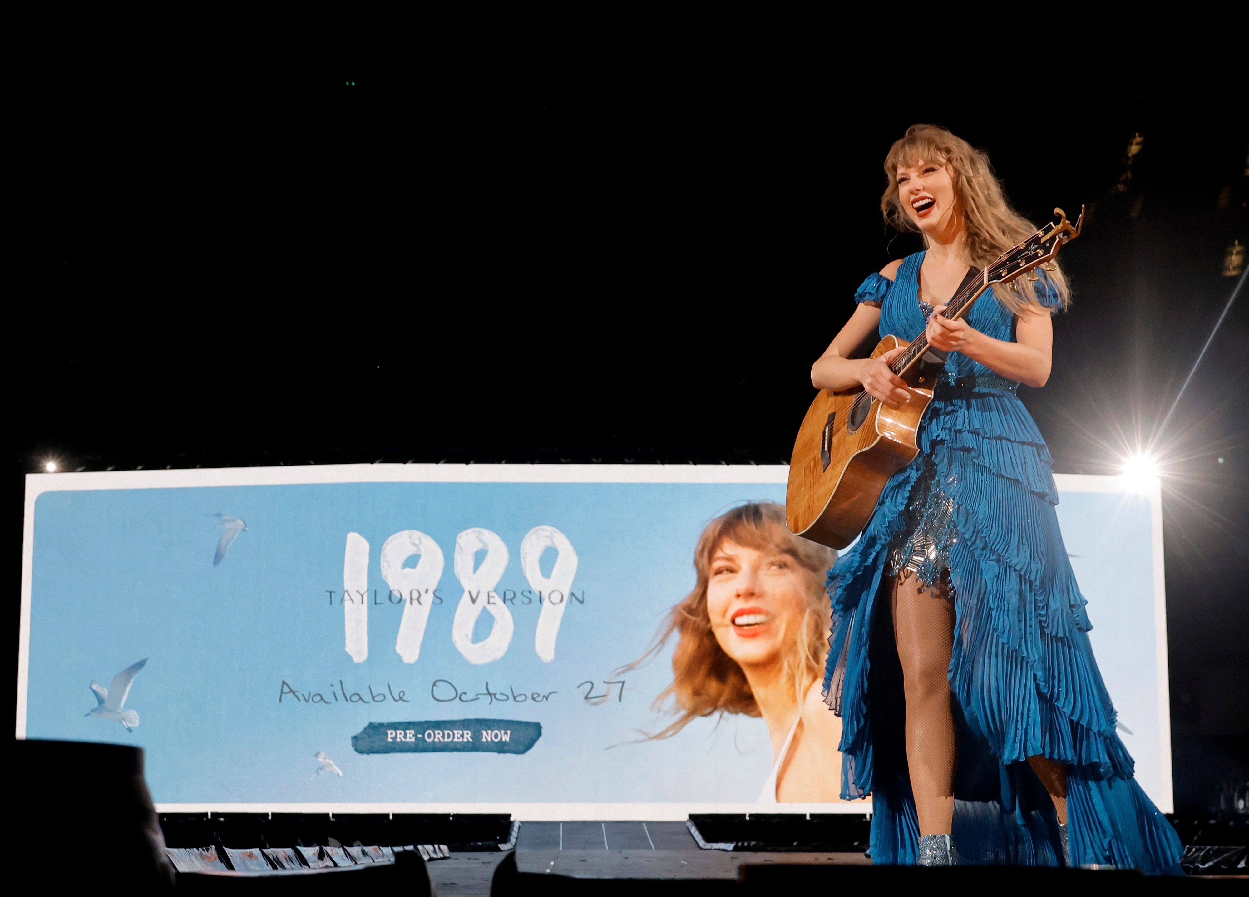 Taylor onstage holding a guitar with an ad for &quot;1989 (Taylor&#x27;s Version)&quot; behind her