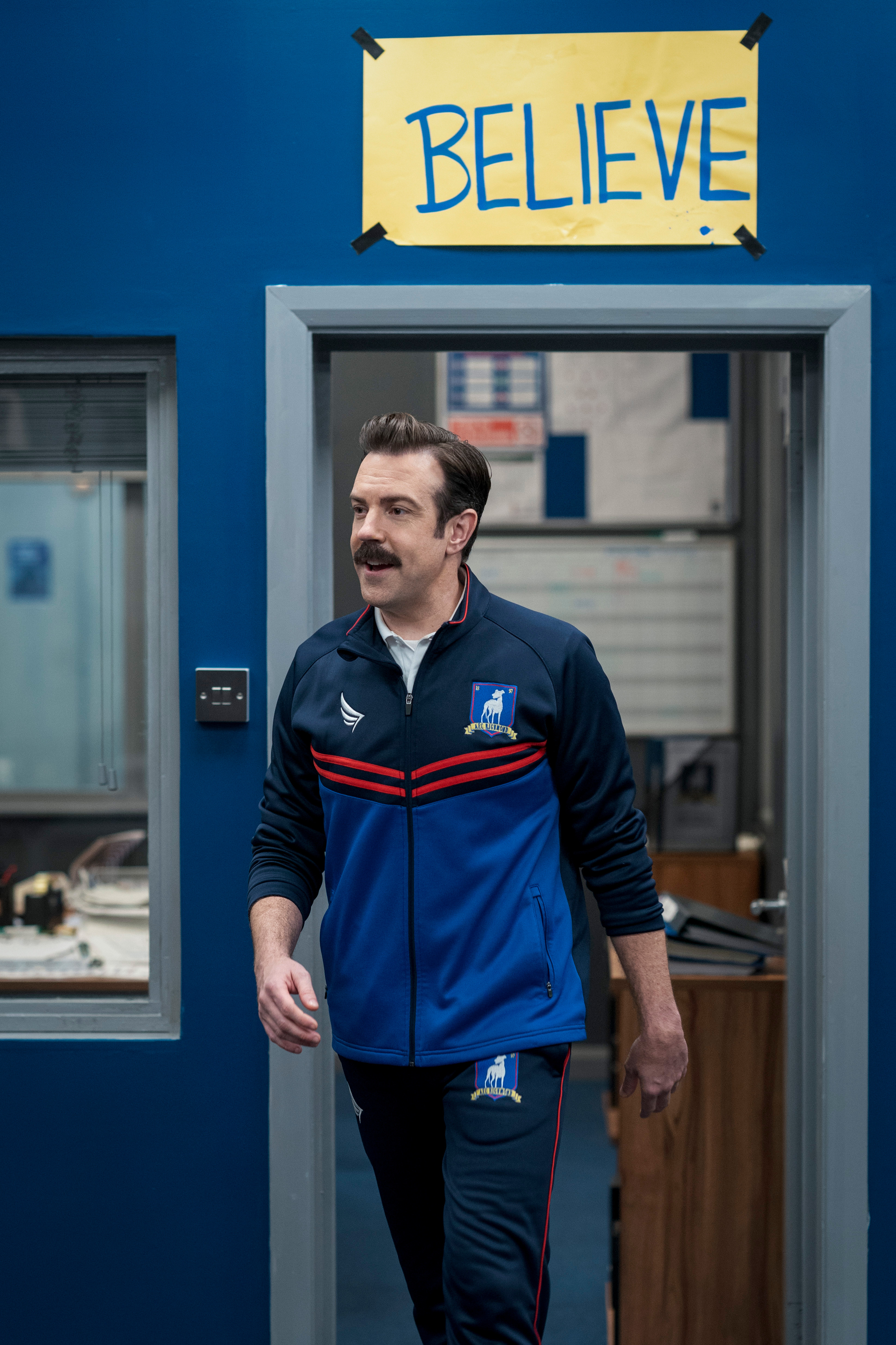 Ted Lasso walking through a door with a sign taped above it that says, "Believe"