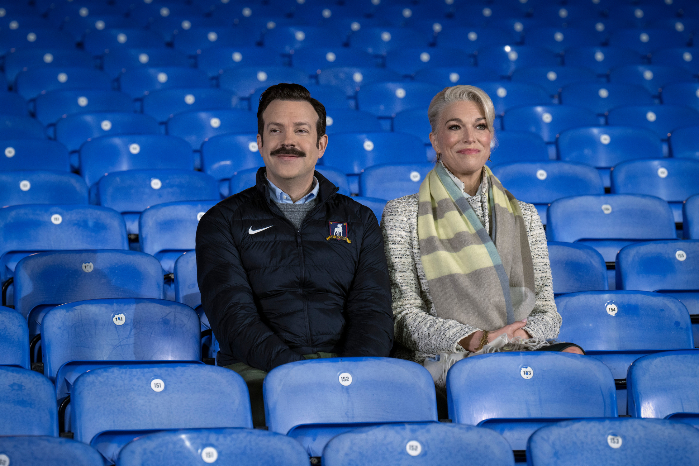Jason Sudeikis as Ted Lassi sitting in the empty stands next to Hannah Waddingham as Rebecca Welton in a scene from  "Ted Lasso"