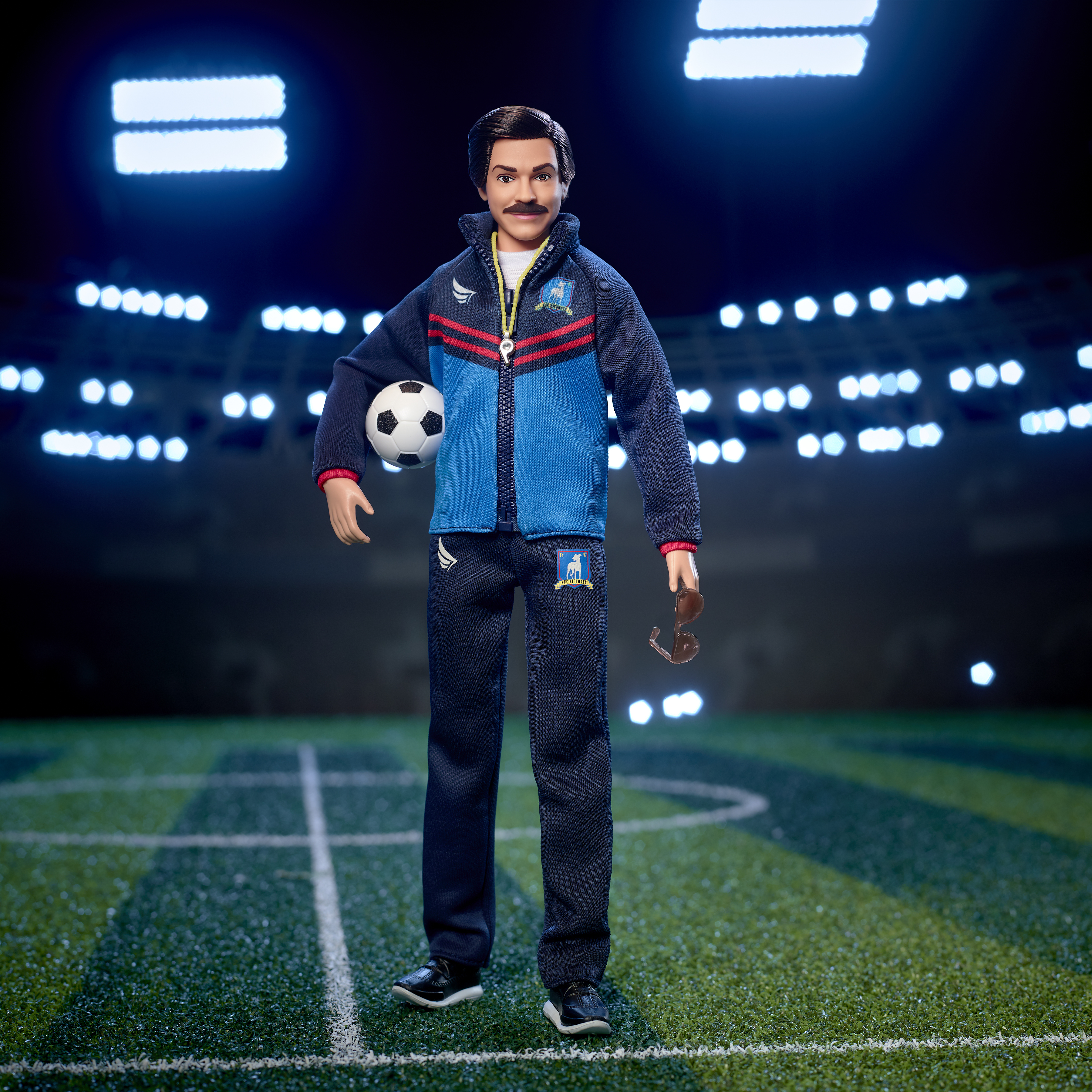 Ted Lasso Barbie doll stands on a soccer pitch holding a soccer ball under one arm and a pair of sunglasses in one hand