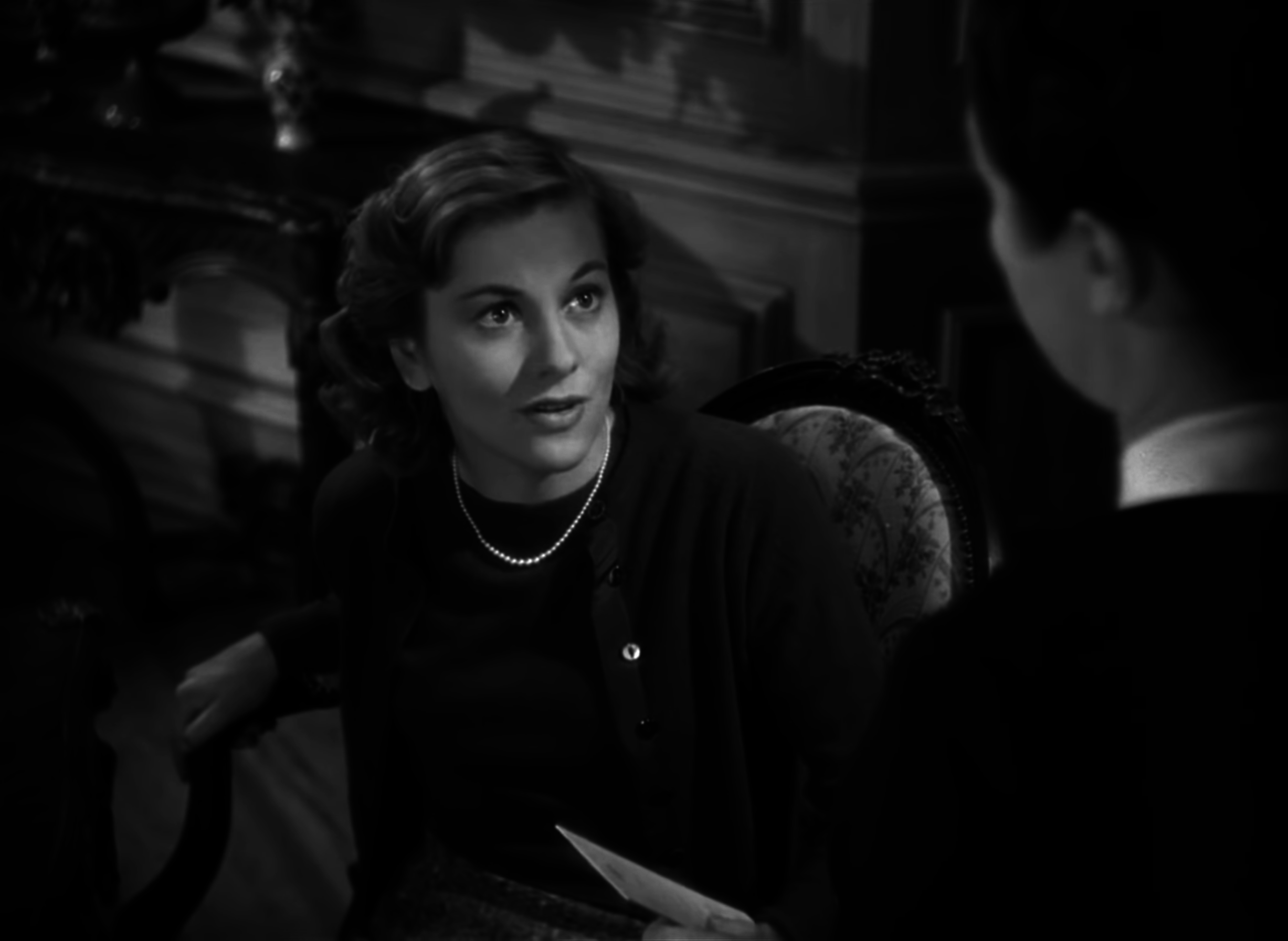 A young Joan Fontaine as Mrs de Winter sitting in a chair looks up at another woman standing over her, whose face we can&#x27;t see