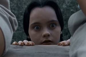 Christina Ricci as Wednesday Addams in 1993's "Addams Family Values." She has wide eyes and a frowning mouth, and is looking over a bench. She is hiding from someone.