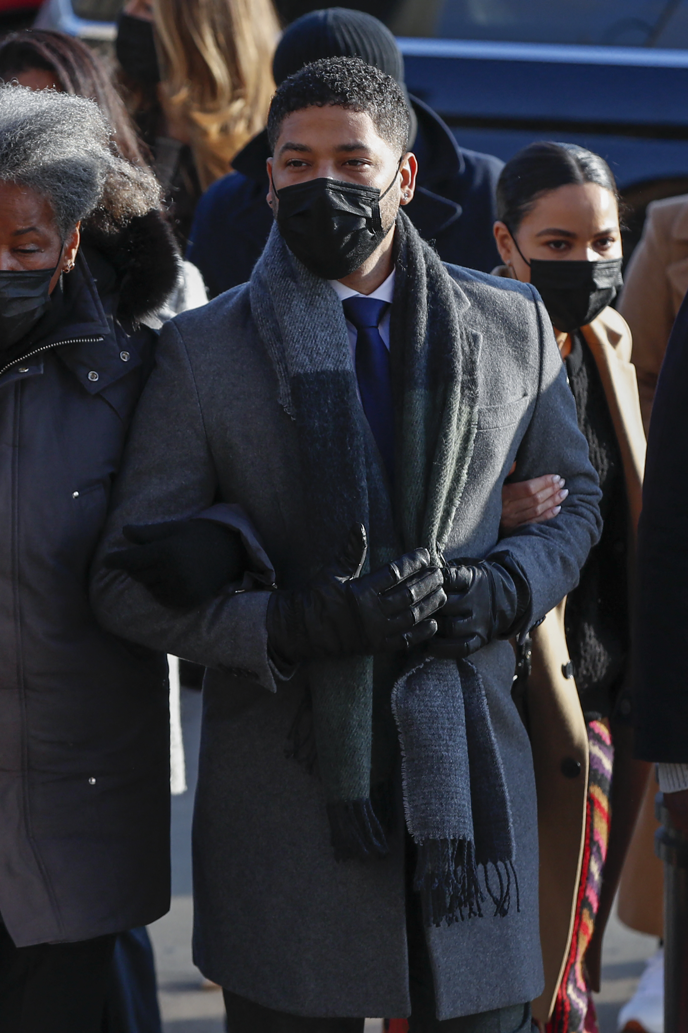 Close-up of Jussie outside in a coat and scarf and wearing a mask and arm in arm with other people
