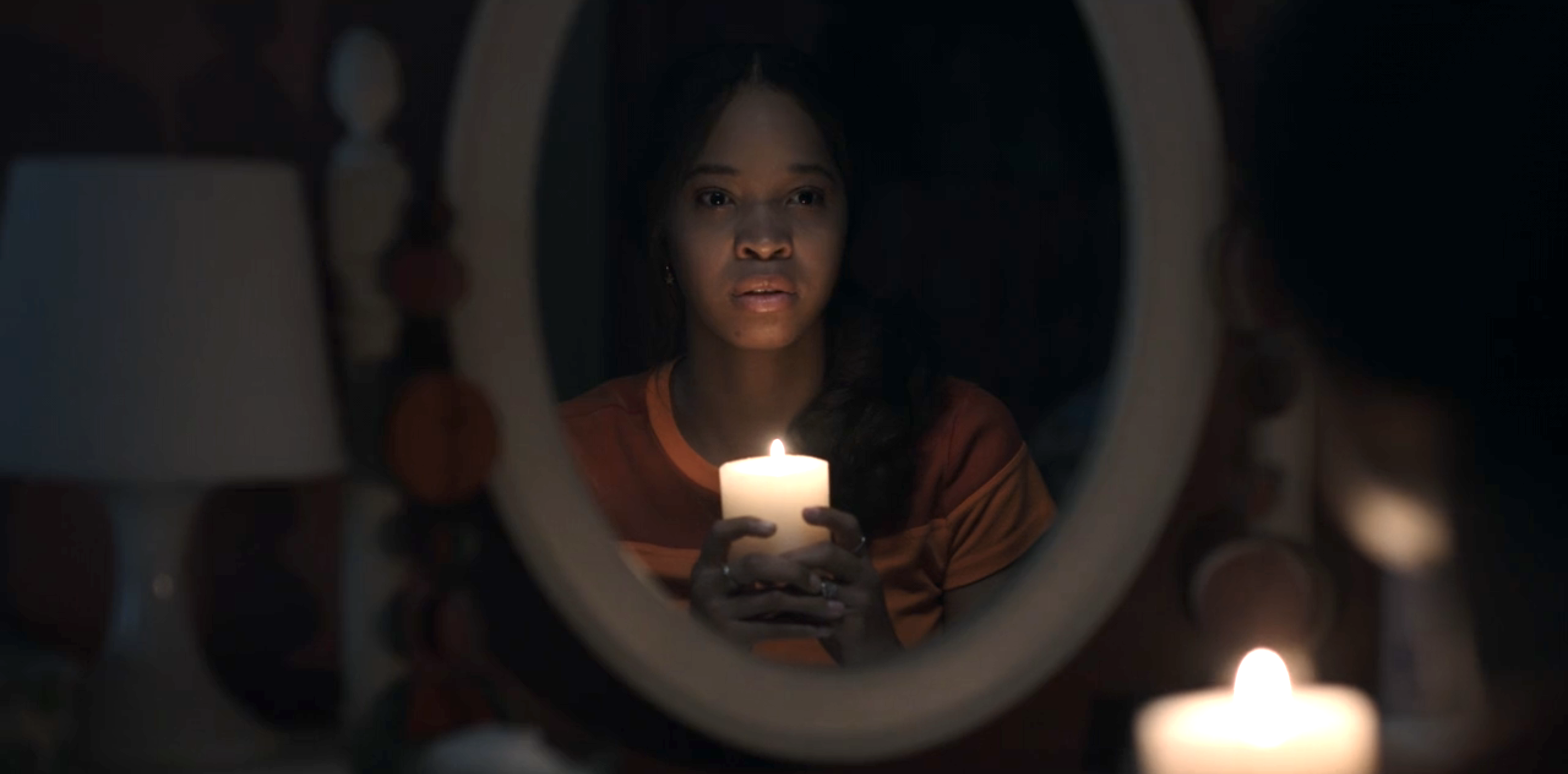 A young woman holding a candle with a very scared expression and looking into the mirror