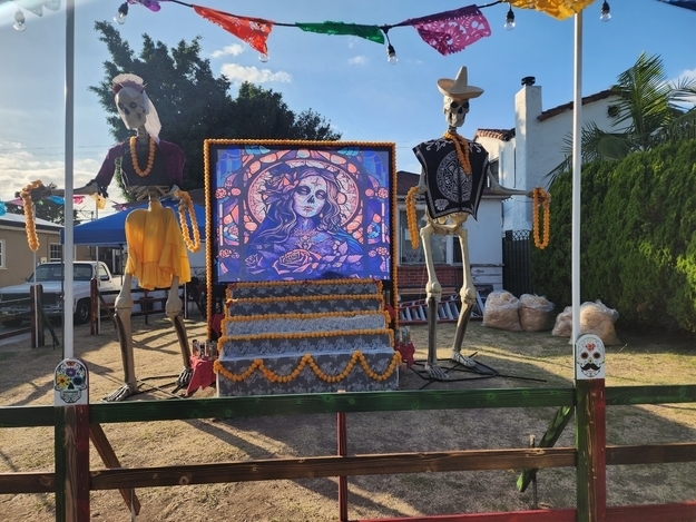 Two skeletons in a yard wearing traditional attire, including hats, with a Catrina painting at the top of steps in the center