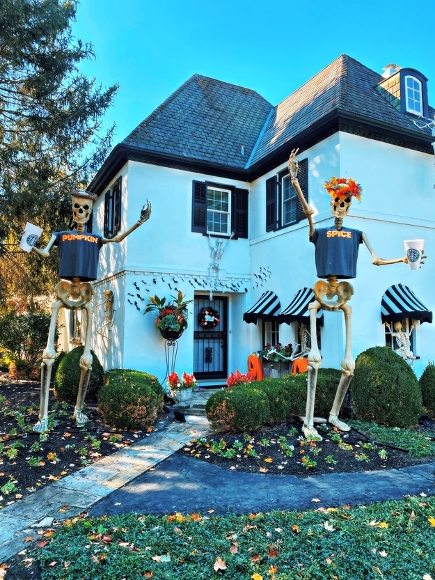 Two skeletons wearing &quot;Pumpkin&quot; and &quot;Spice&quot; T-shirts and holding large coffee cups in front of a house