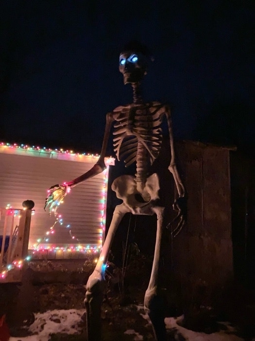 A skeleton with lit eyes holding Christmas lights that are also on a window at night