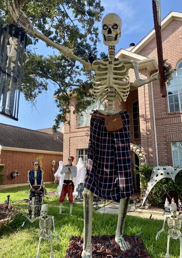 A skeleton in front of a building with a fanny pack and wearing a skirt, and holding what looks like a tennis racket, with other skeletons behind it