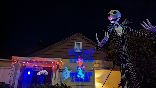 A skeleton in a pin-striped suit in front of a house at night with a &quot;Trick or Treat&quot; message reflecting on the house