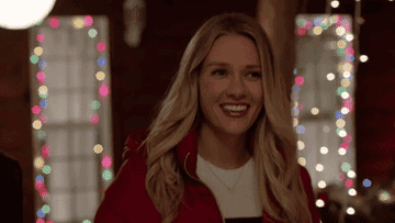 a gif of a blond actress smiling and laughing with Christmas lights in the background