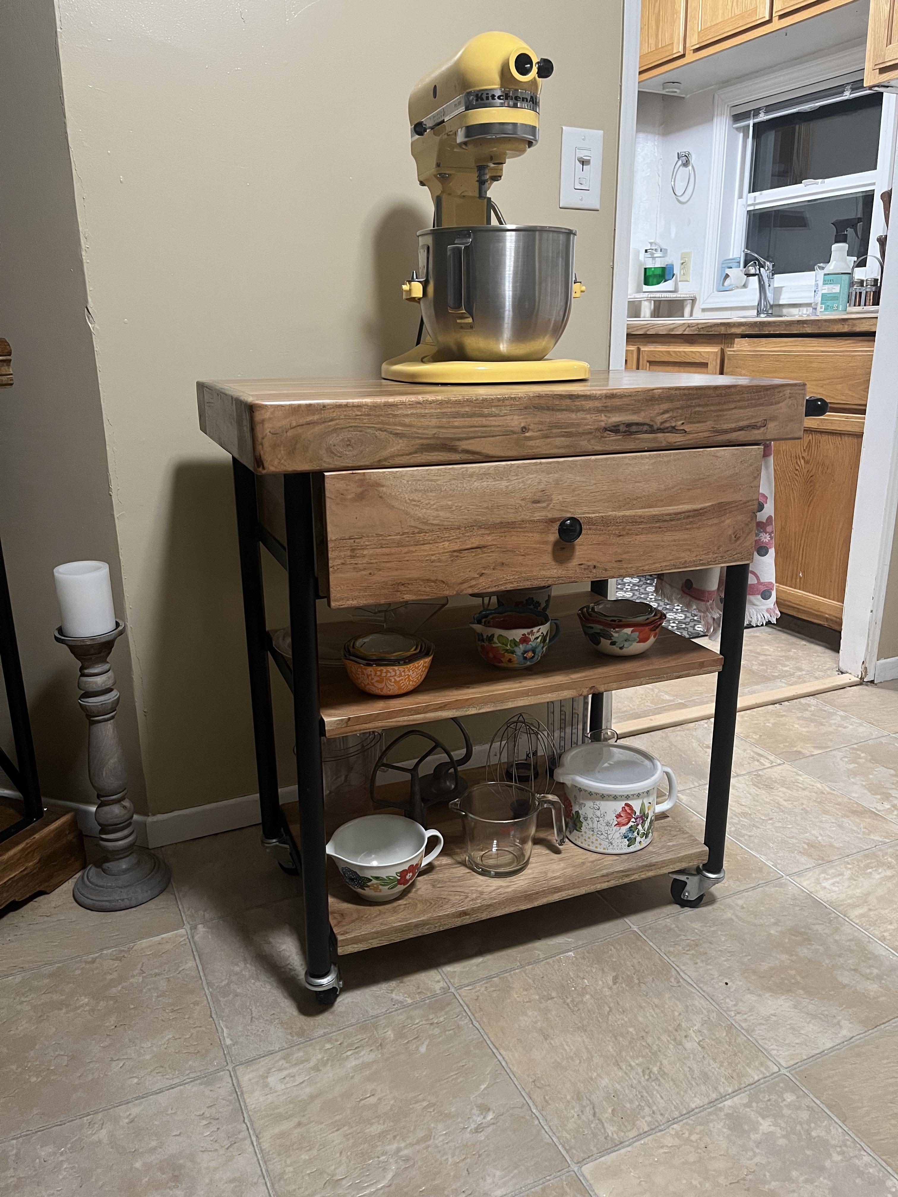 A mixer on top of a wood kitchen cart with various kitchen and baking supplies underneath