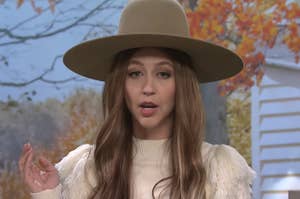 Heidi Gardner wearing a large hat and sweater and standing in front of an autumn tree in an SNL sketch