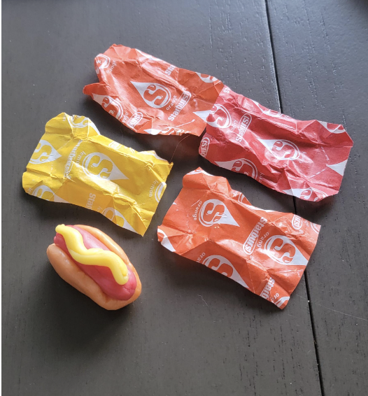 A hot dog (with bun and mustard) made out of Starbursts candies next to four Starbursts candy wrappers of matching colors