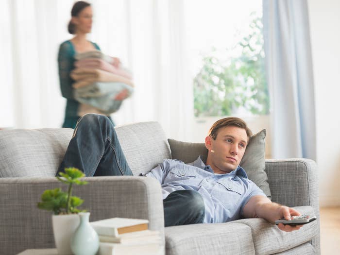 guy watching tv on the sofa while a woman carries laundry behind him