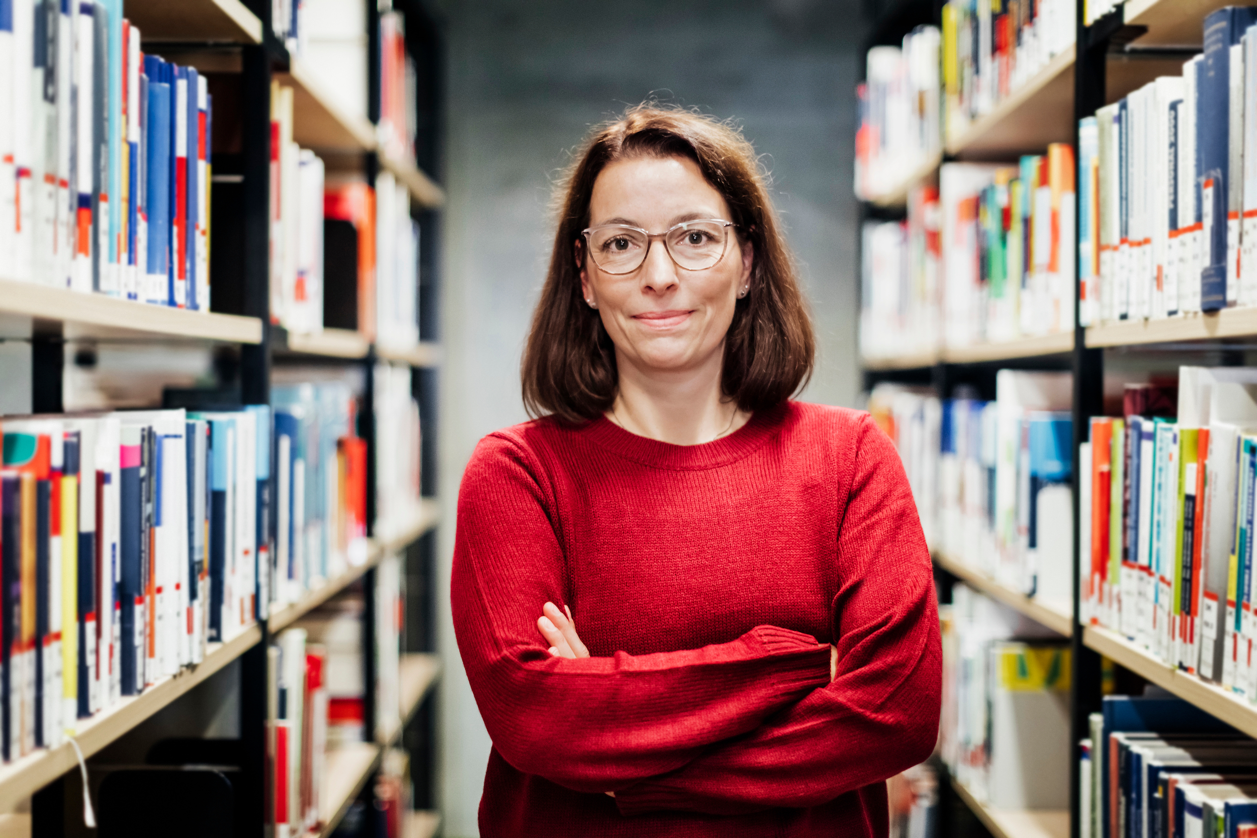 A woman in a library wearing a red sweater with her arms crossed