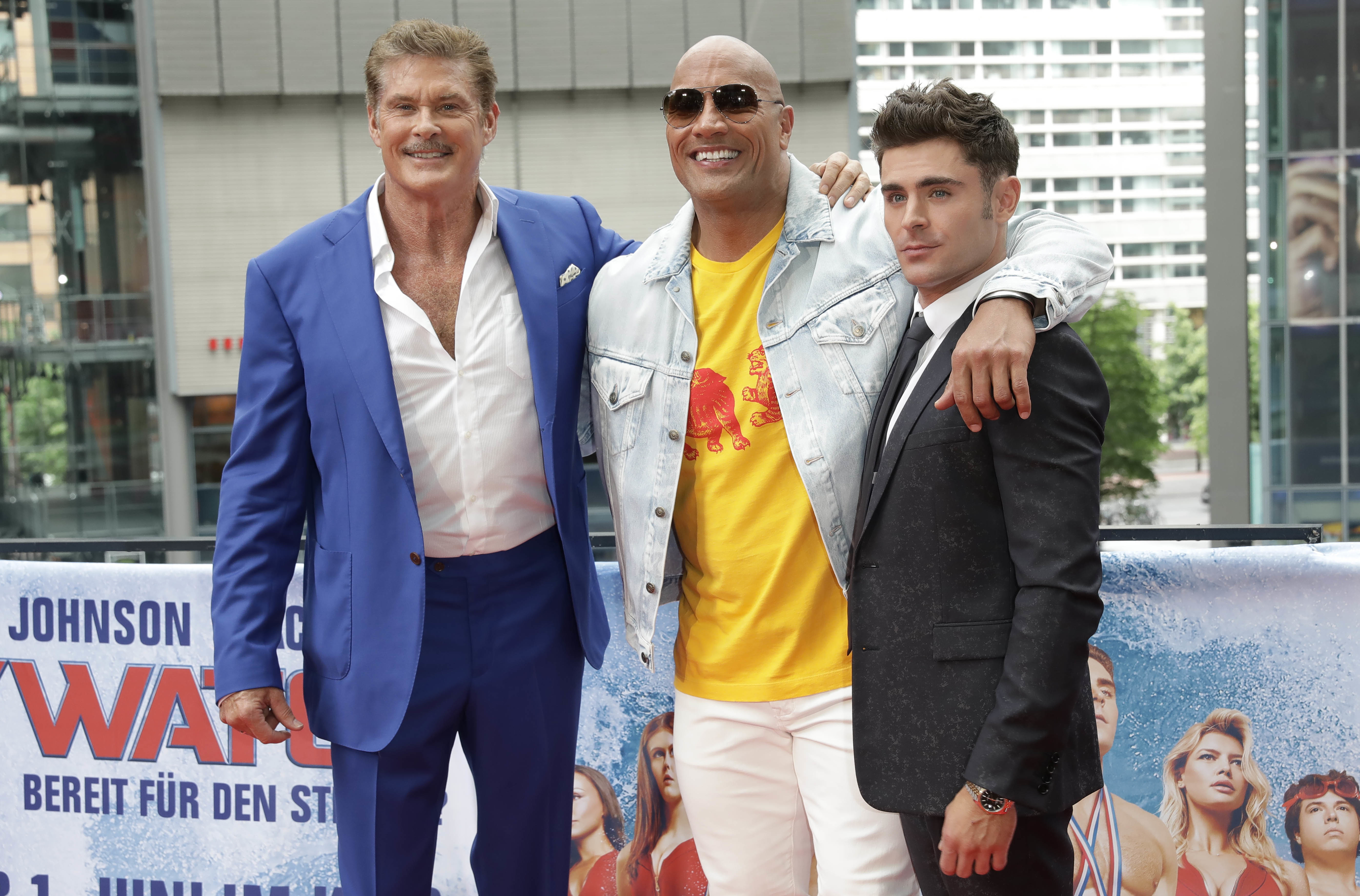 Close-up of Zac smiling and standing with Dwayne Johnson and David Hasselhoff at a media event