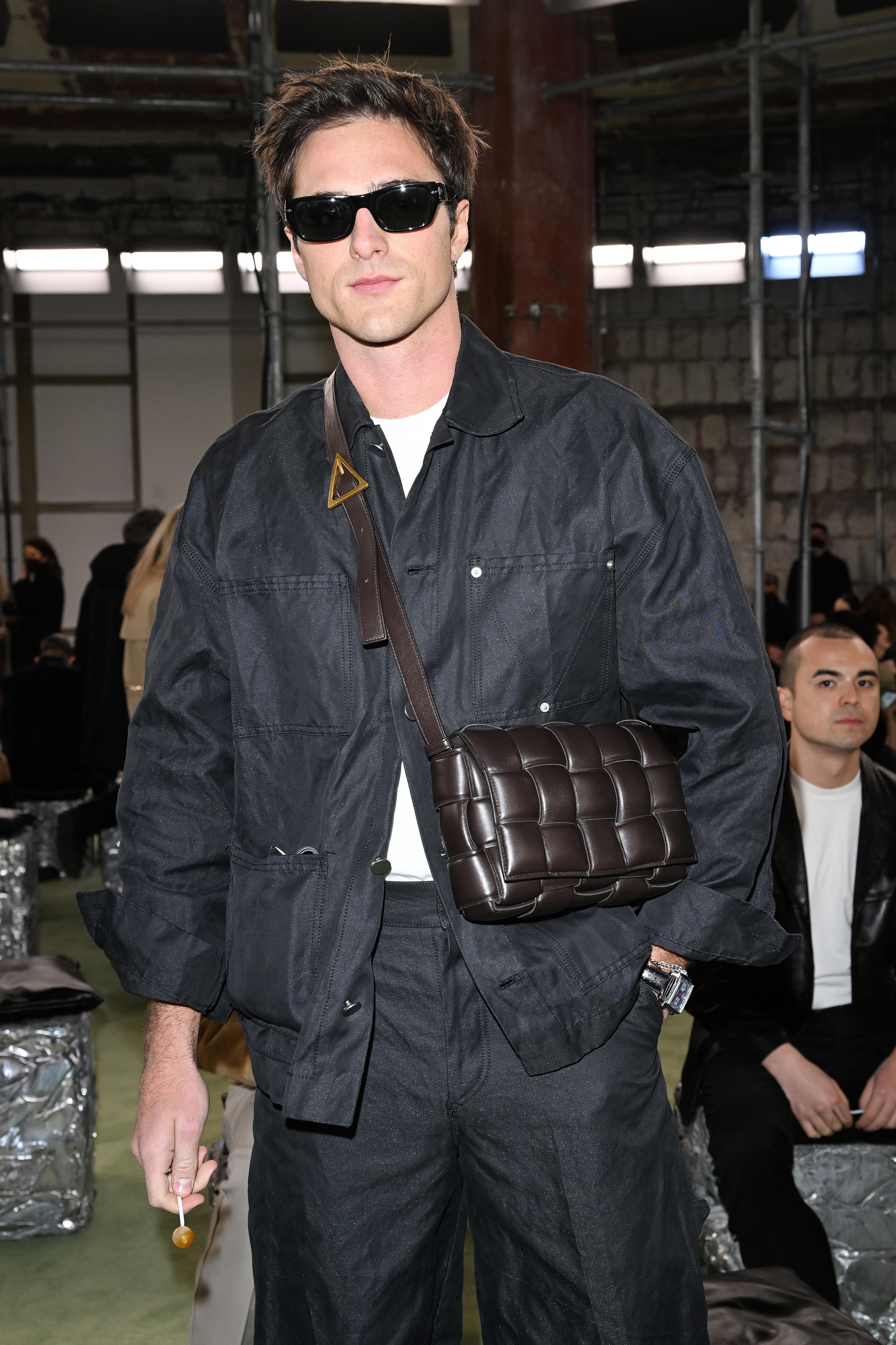 Close-up of Jacob in a jacket with a small shoulder bag that reaches his waist and holding a lollipop