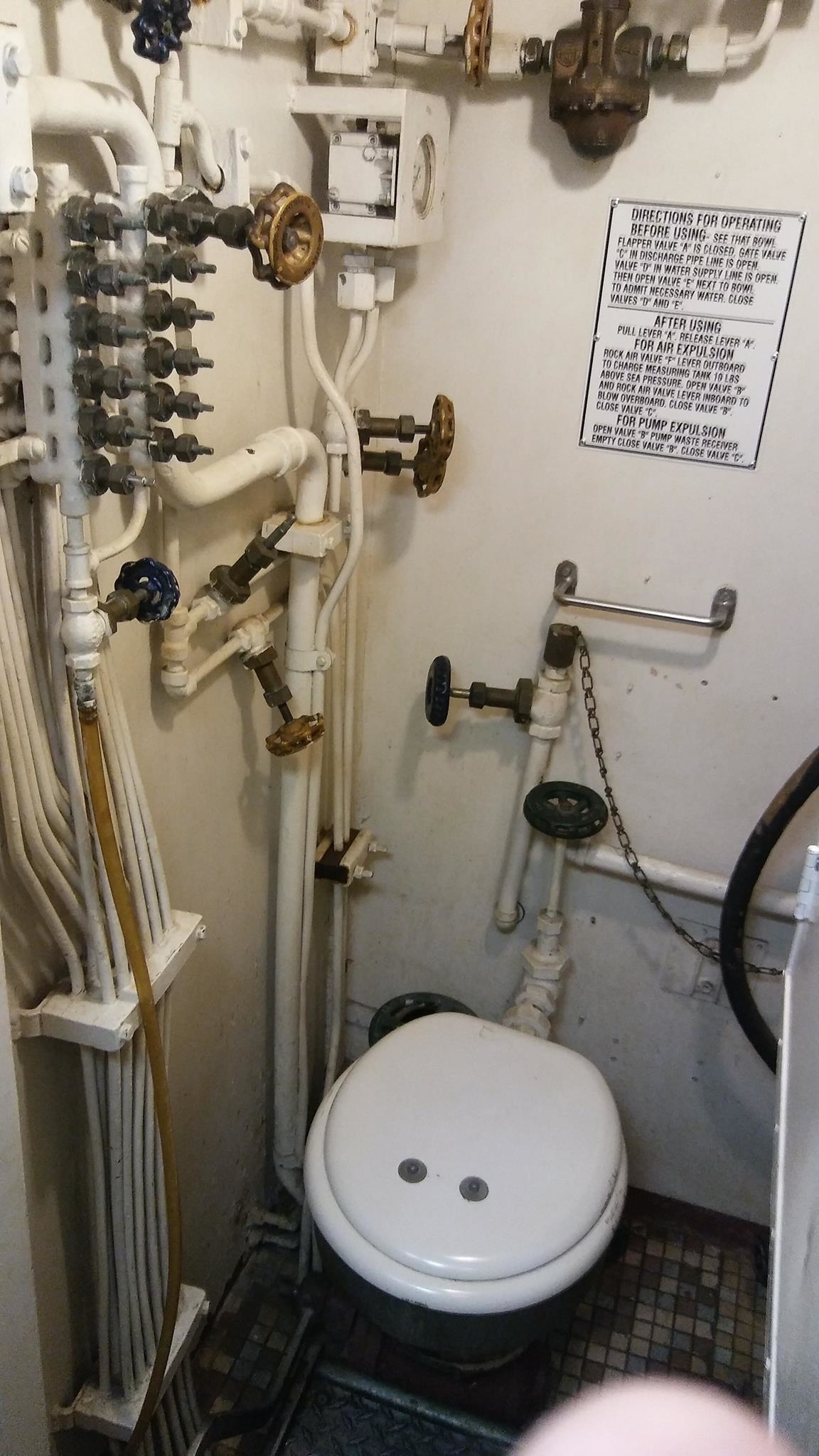 A toilet bowl surrounded by pipes and chains and cords and valves and knobs, with a sign with lots of text labeled, &quot;Directions for operating: before and after using&quot;