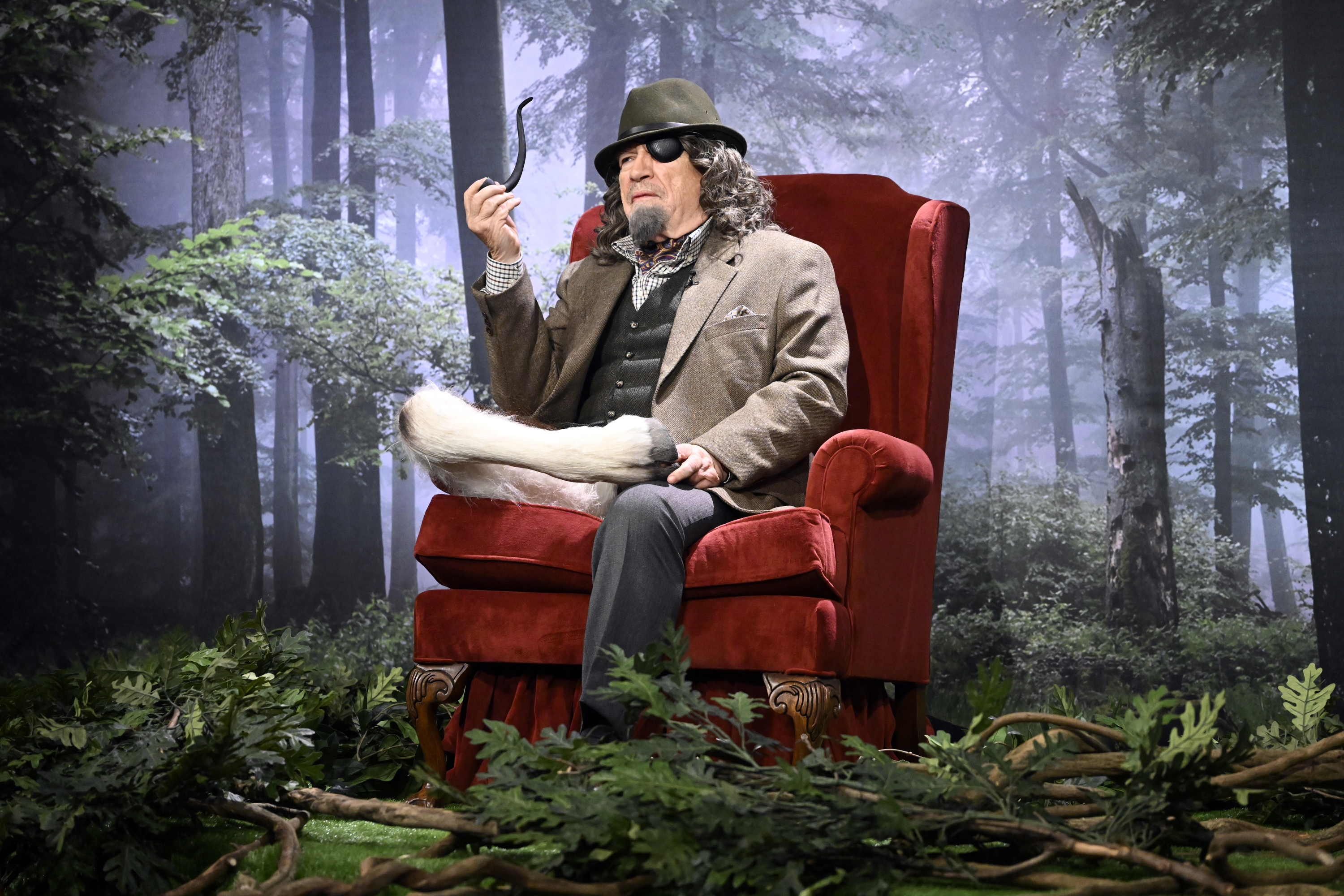 brian cox sitting on an armchair on a tv set made to resemble a forest. one of his legs is a goat&#x27;s leg. he wears a wig, fake beard, and fedora along with an eyepatch, and smokes a pipe