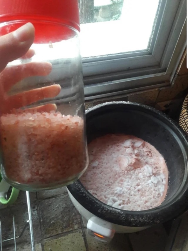 Someone holding up a container of Himalayan pink salt with a rice cooker full of pinkish salt broth in the background