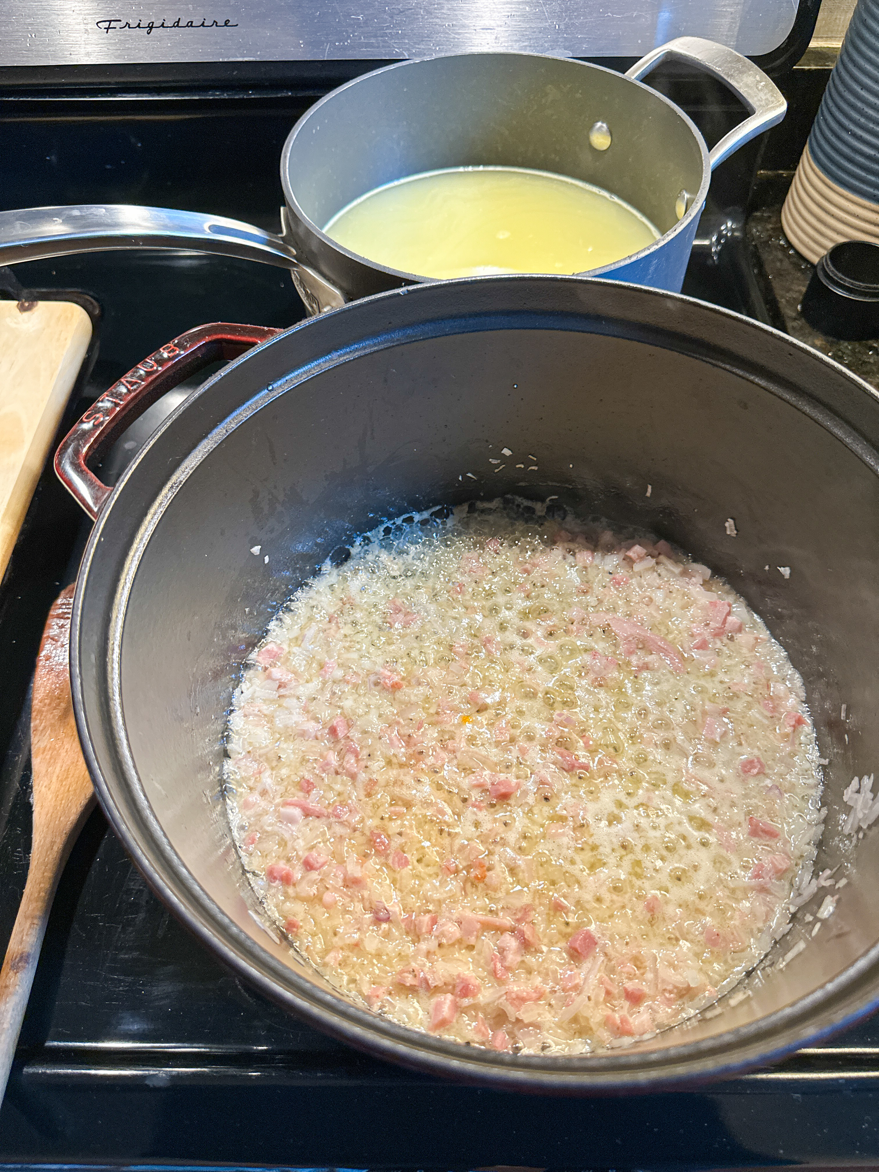 Butter, pancetta, and shallot sautéing in a pot with a saucepan of stock on the back burner