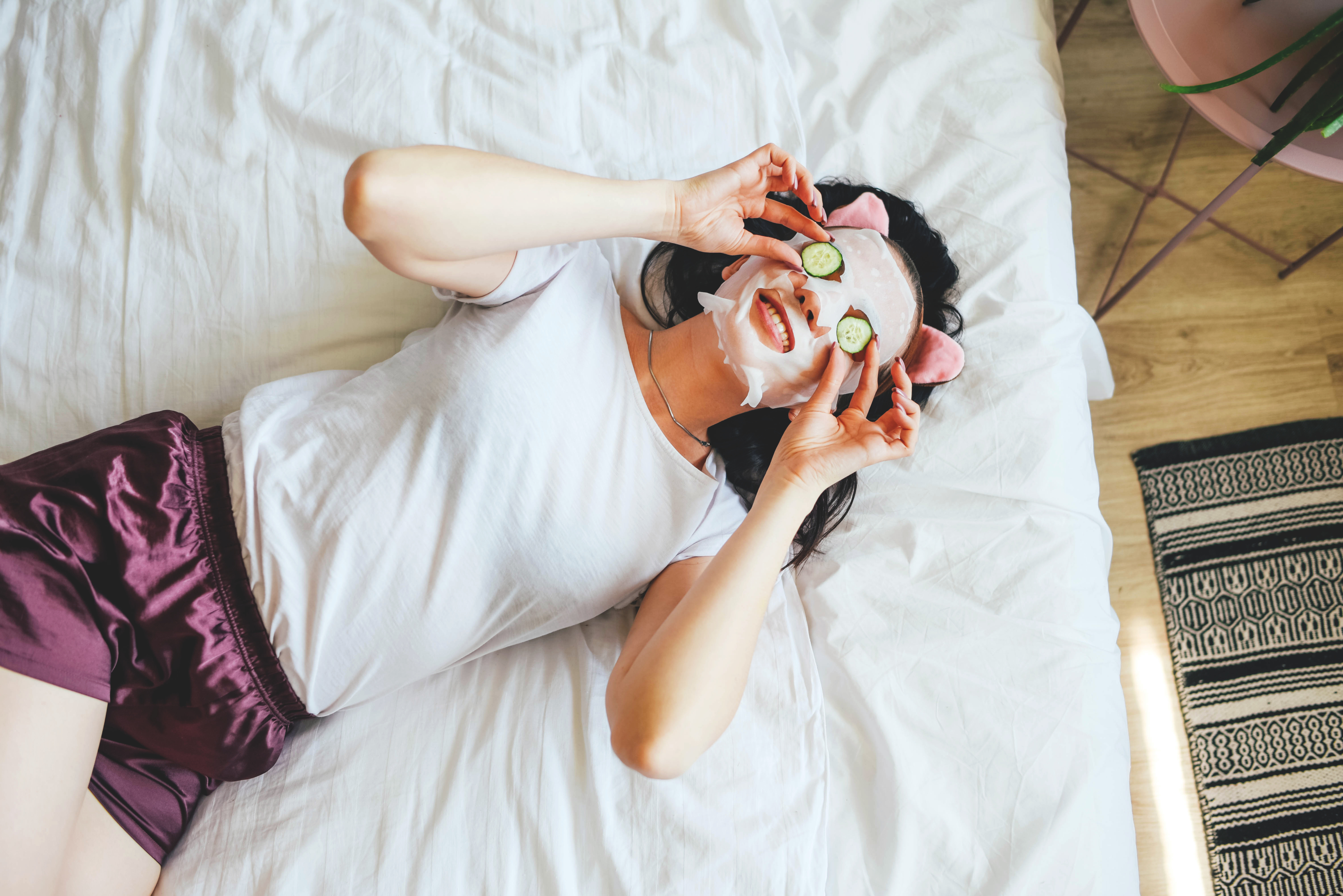 A woman with a face mask on and cucumber slices on her eyes while lying in bed