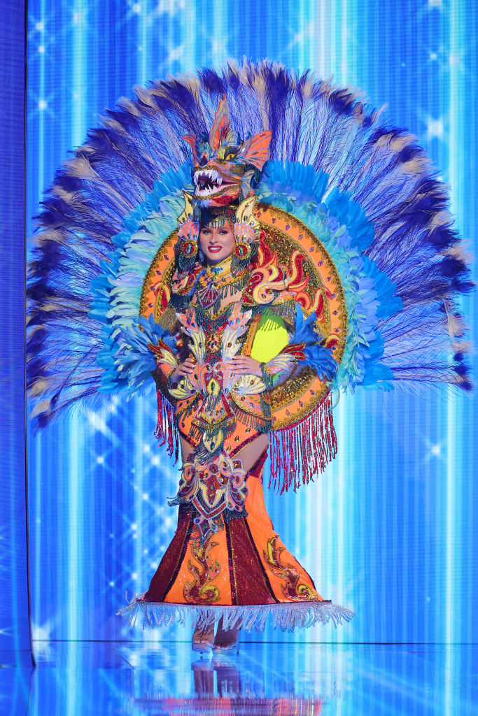 She&#x27;s wearing a colorful, ornate, bejeweled outfit with peacock-like, feathery train and dragon headpiece