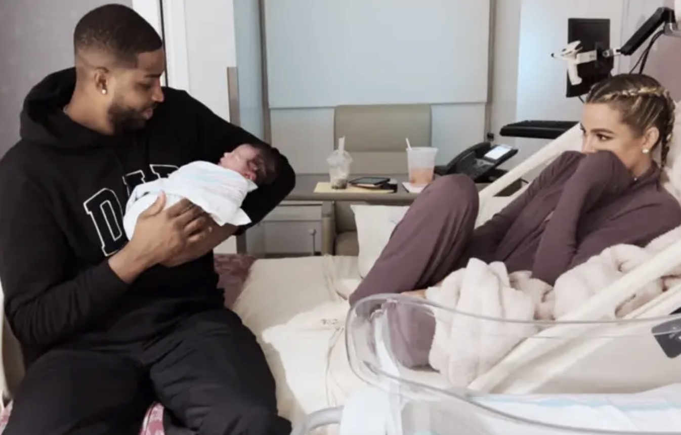 Close-up of Tristan holding a baby and sitting on a bed with Khloé