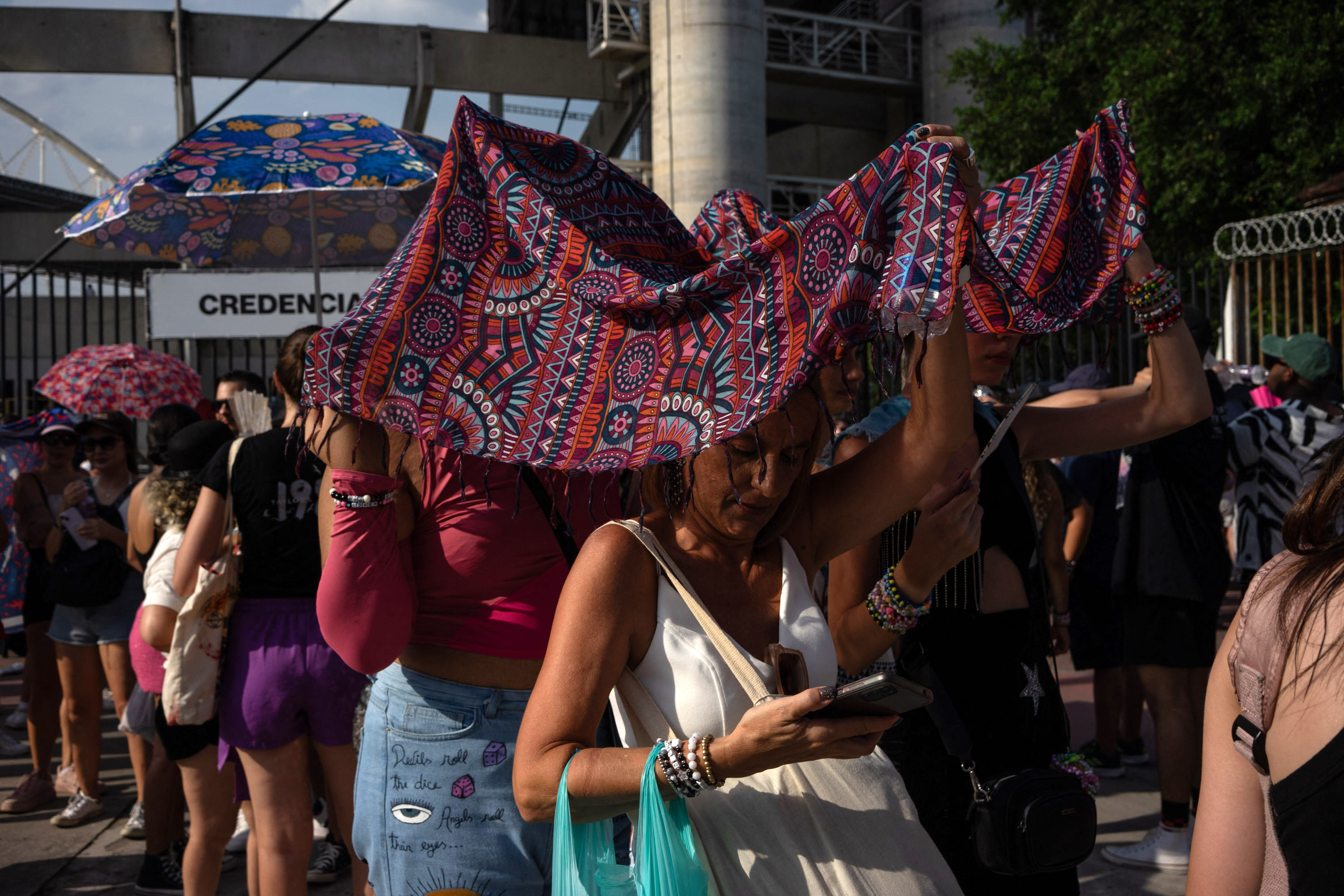 Fans standing in line, some holding fabrics or umbrellas over their heads