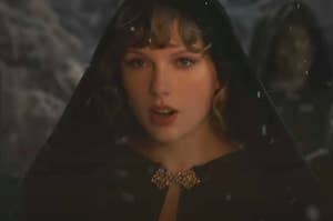 Taylor Swift walking in a forest while wearing a cloak as snow falls around her in the Willow music video