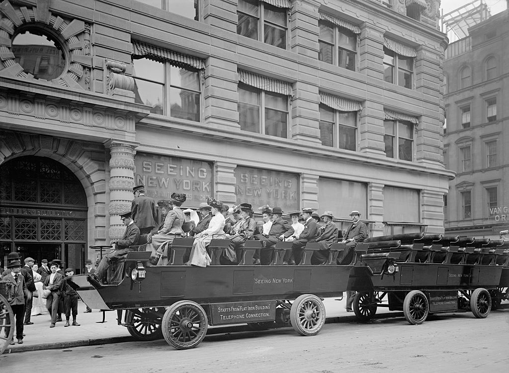 An old NYC tour bus with women and men in long dresses sitting on a raised platform on wheels with no sides