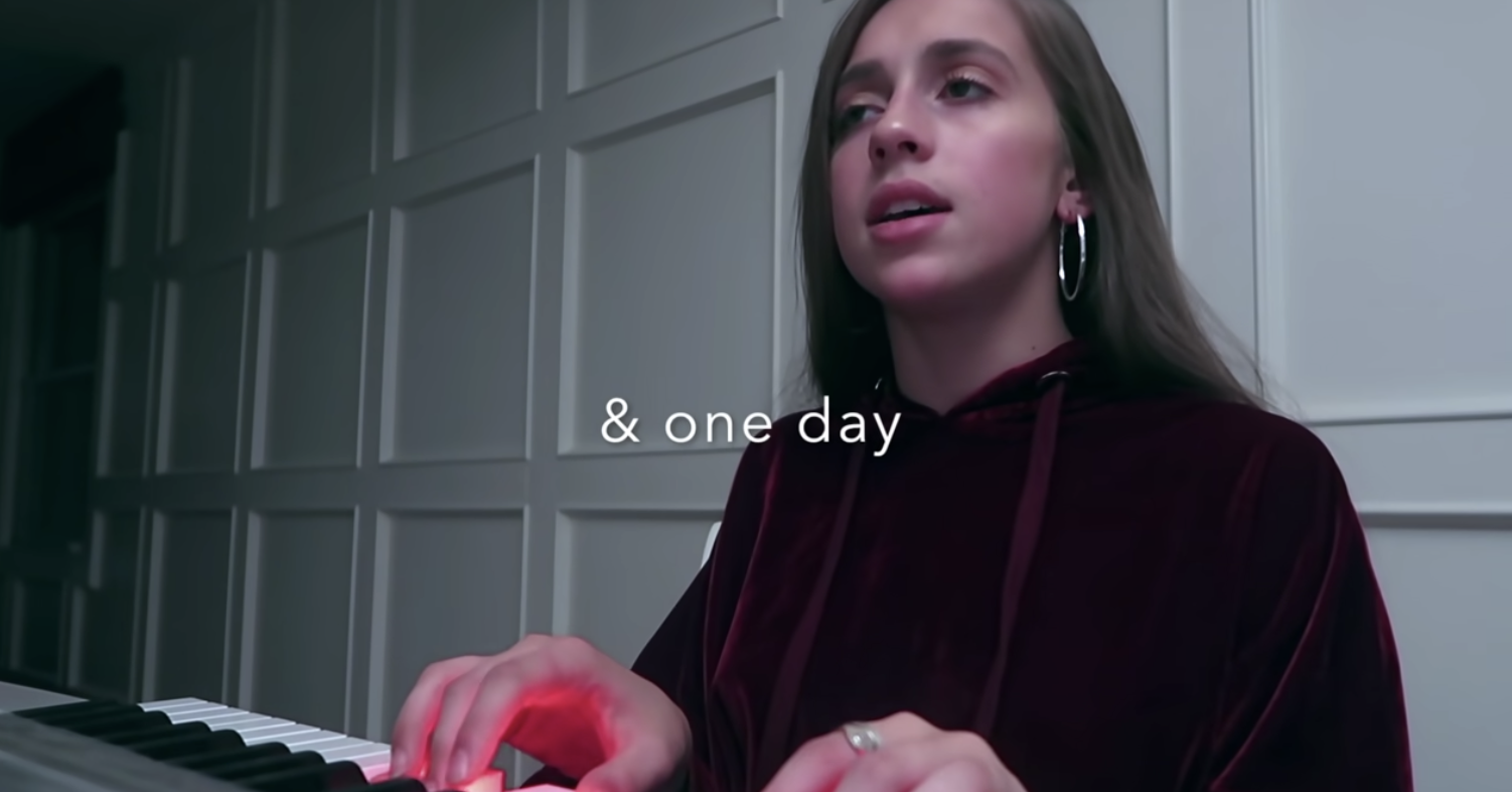 Screenshot of Tate Mcrae&#x27;s video for the original song &quot;one day.&quot; She sits and sings at a piano.