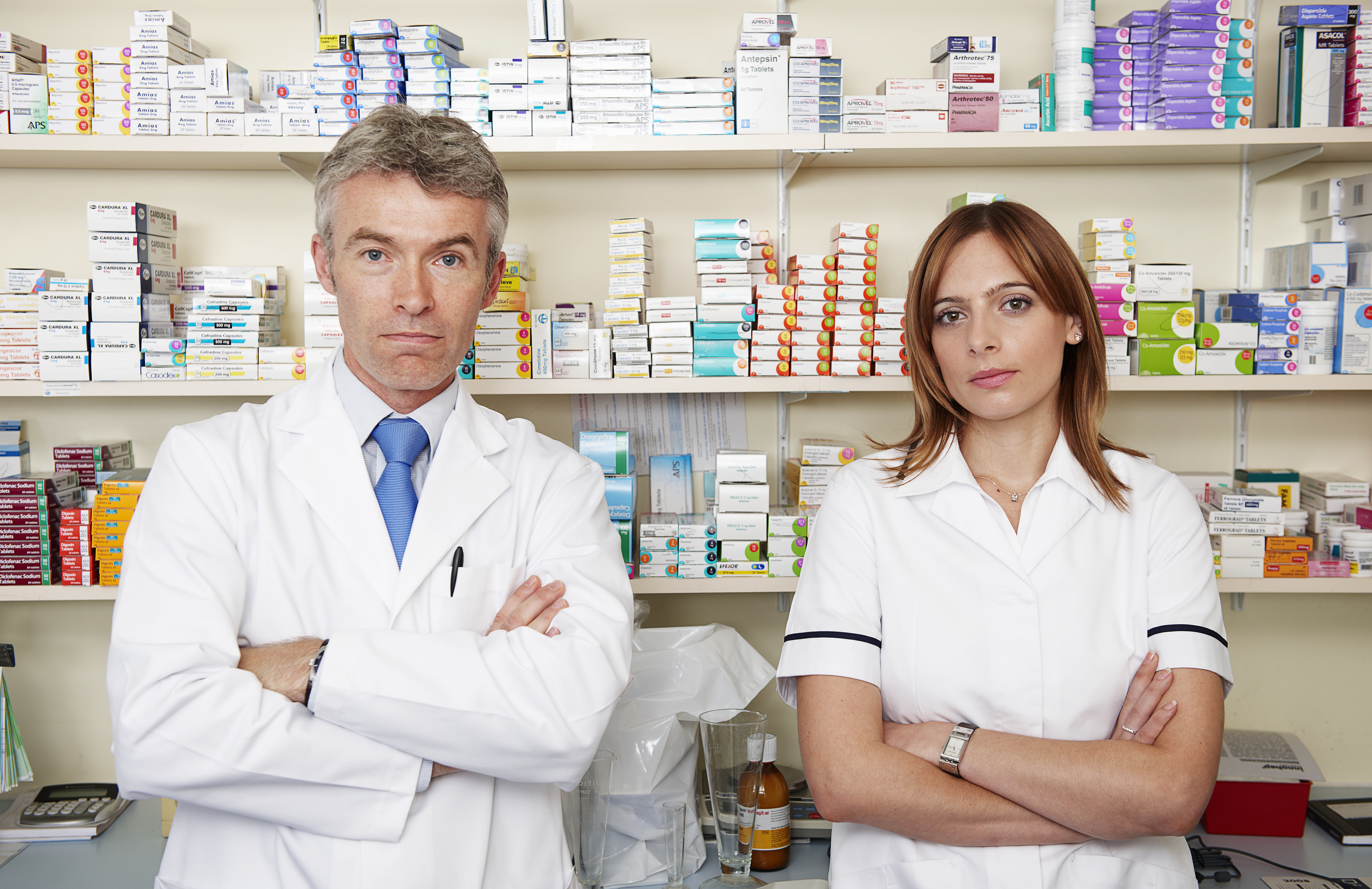 Two pharmacists with their arms crossed as they stand in front of shelves with medicine