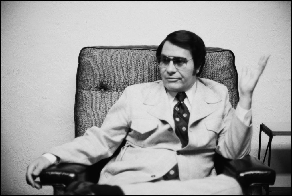 Jim Jones sitting in a chair wearing sunglasses and a leisure suit