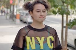 Ilana from Broad City walking down the street wearing a mesh top that says NYC