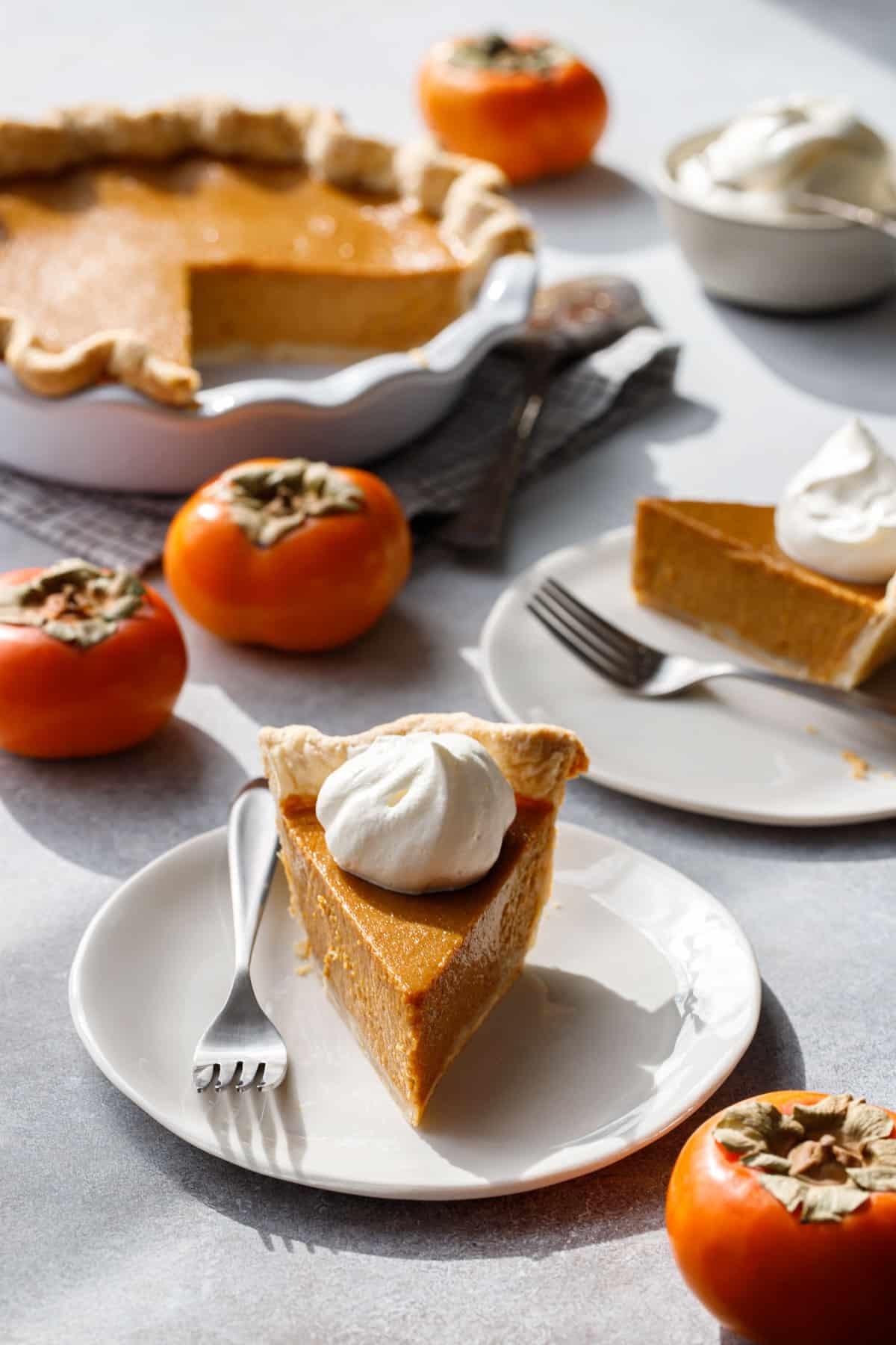 A slice of persimmon pie topped with whipped cream and surrounded by whole persimmons