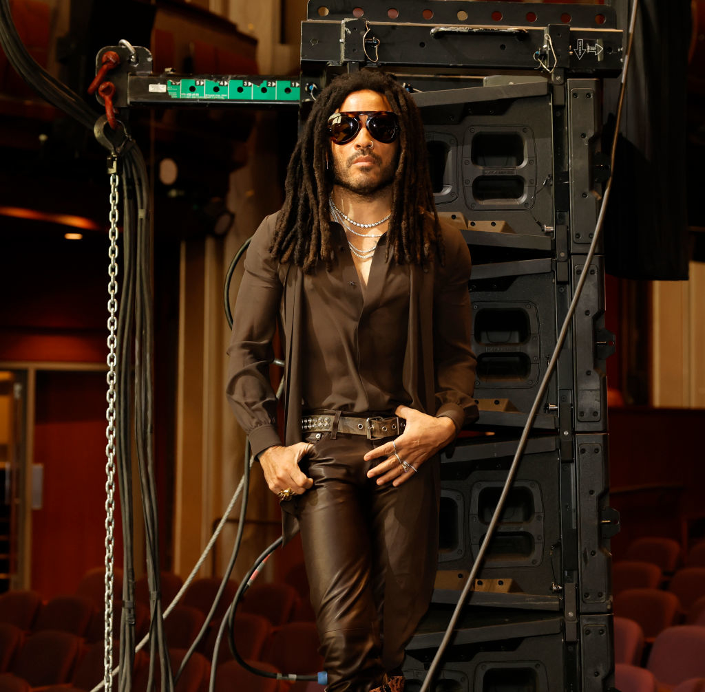 Lenny posing against machinery for a photoshoot wearing leather pants and a sheer button-down