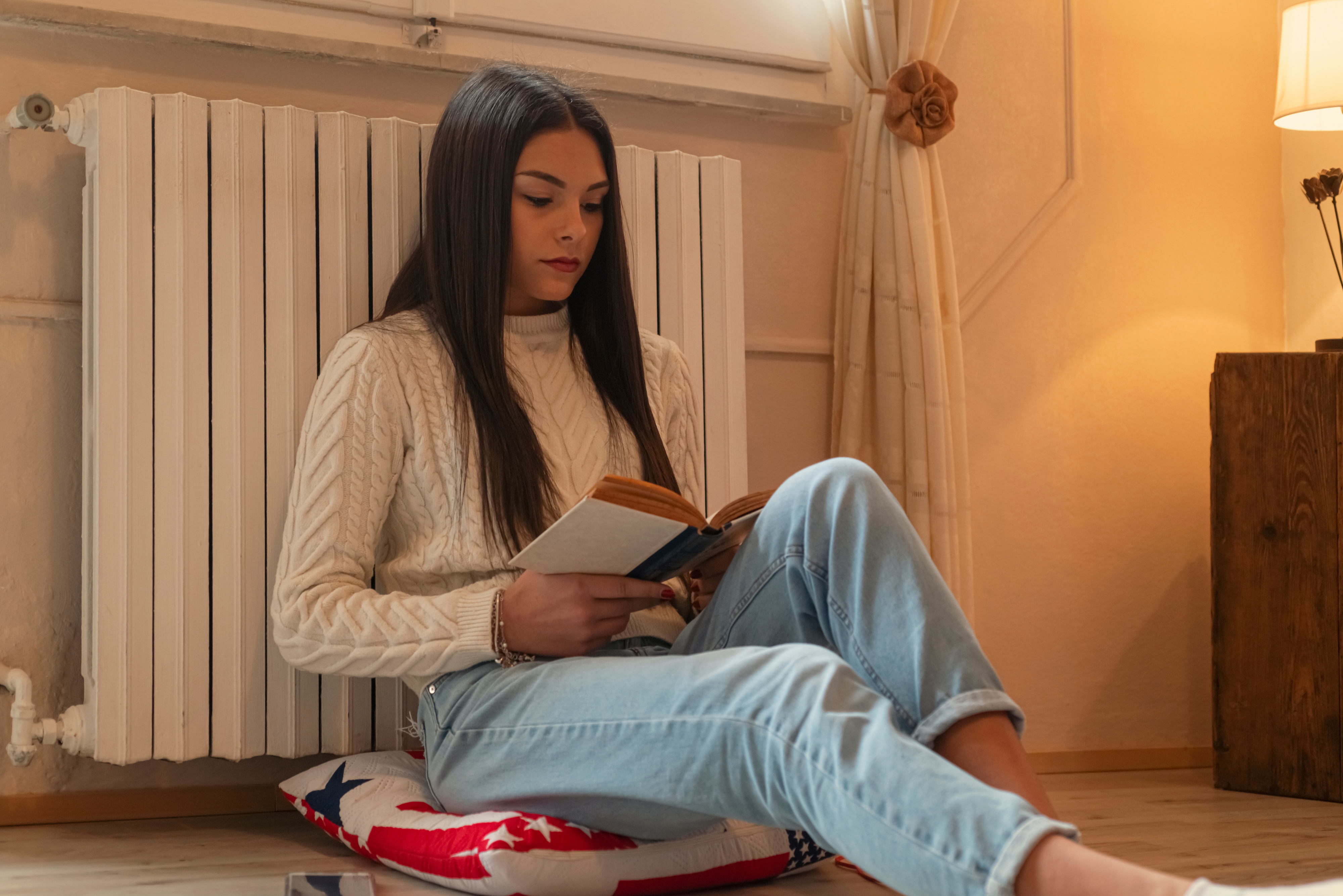 a young woman in a sweater and jeans sits in her dorm room reading a book
